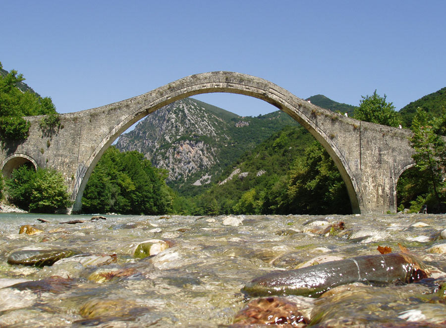 Το ιστορικό γεφύρι της Πλάκας στην Ήπειρο, πάνω από πετρώδες ποτάμι, με βουνά στο βάθος.
